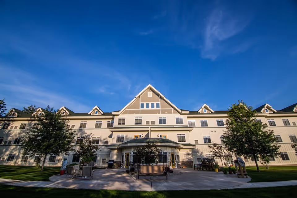 Front exterior view of a large, multi-story assisted living facility building with a peaked roof and multiple windows under a clear blue sky. There is a paved patio area with outdoor seating and trees in front of the building.