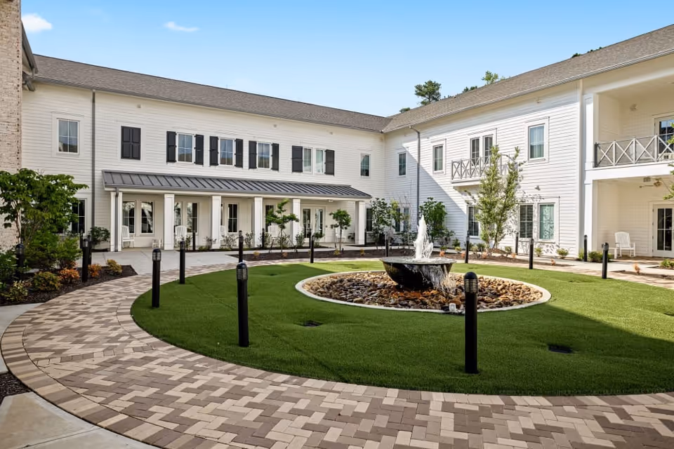 A courtyard area at The Claiborne at Westlake featuring a circular water fountain surrounded by rocks and green grass, with a paved walkway encircling the fountain. The courtyard is bordered by a two-story white building with black shutters, balconies, and a covered porch with seating. Small trees and shrubs are planted around the courtyard.