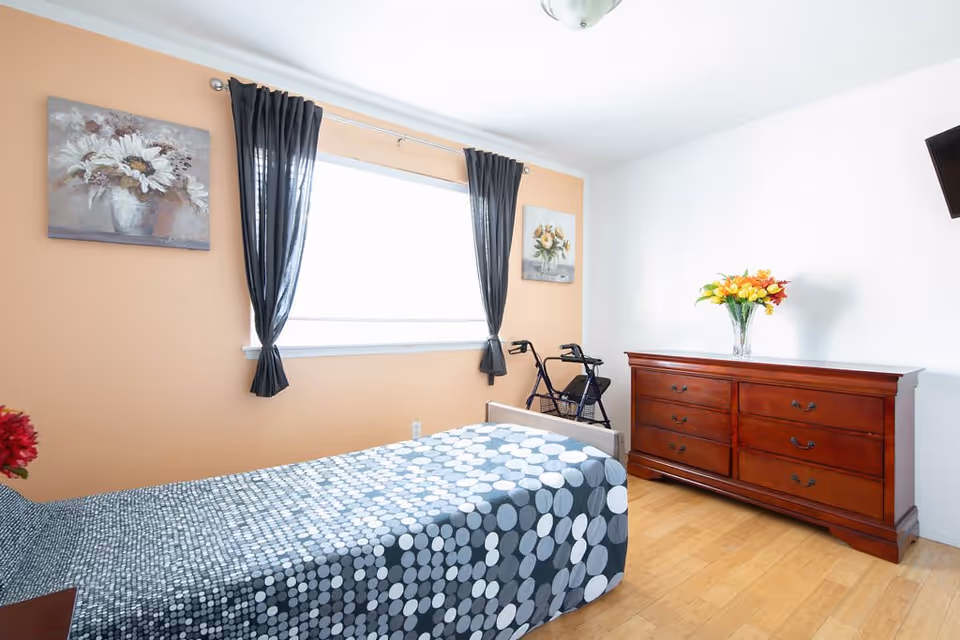 A bright bedroom with a single bed covered in a gray and white polka dot patterned bedspread. There is a wooden dresser with six drawers on the right side, topped with a vase of colorful flowers. A walker is positioned near the window, which has black curtains tied back. Two floral paintings hang on the walls, one above the bed and one next to the window. The floor is light wood, and a flat-screen TV is mounted on the wall above the dresser.