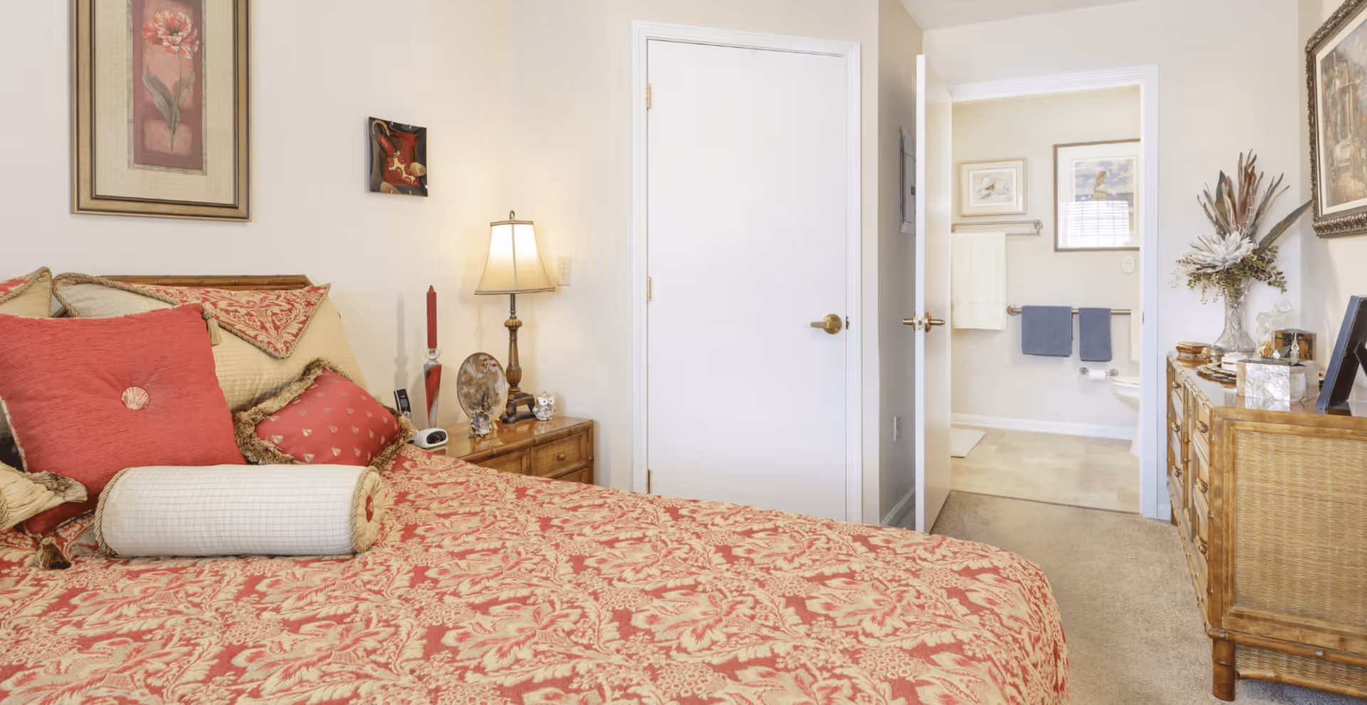 Sunlit bedroom with a red patterned bedspread, nightstand and lamp, and an open door leading to a bathroom.