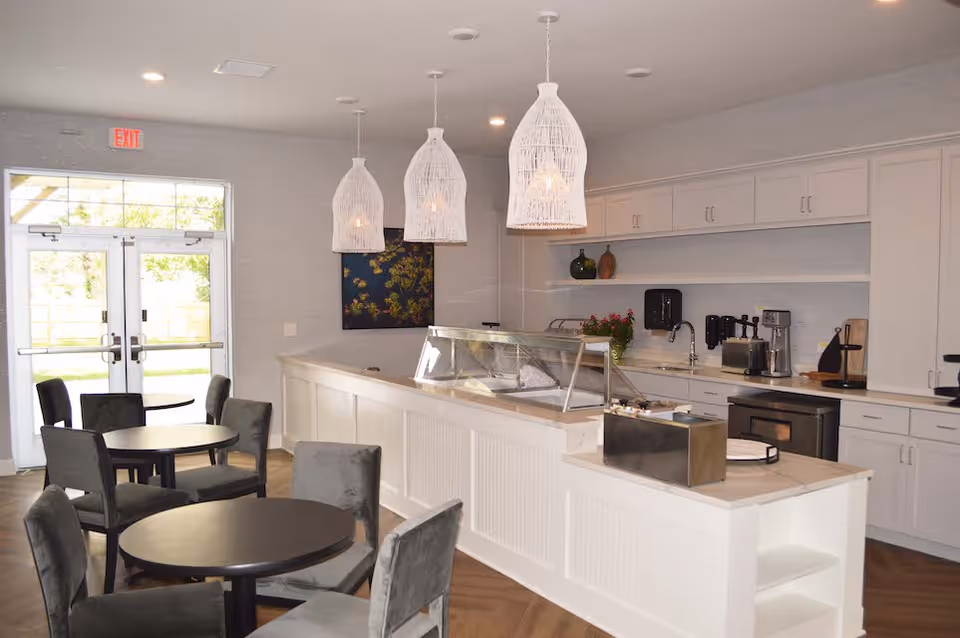 Interior view of a dining area in a senior living facility featuring round tables with gray upholstered chairs, a serving counter with a glass sneeze guard, white cabinetry, pendant lights hanging from the ceiling, and double glass doors leading outside.