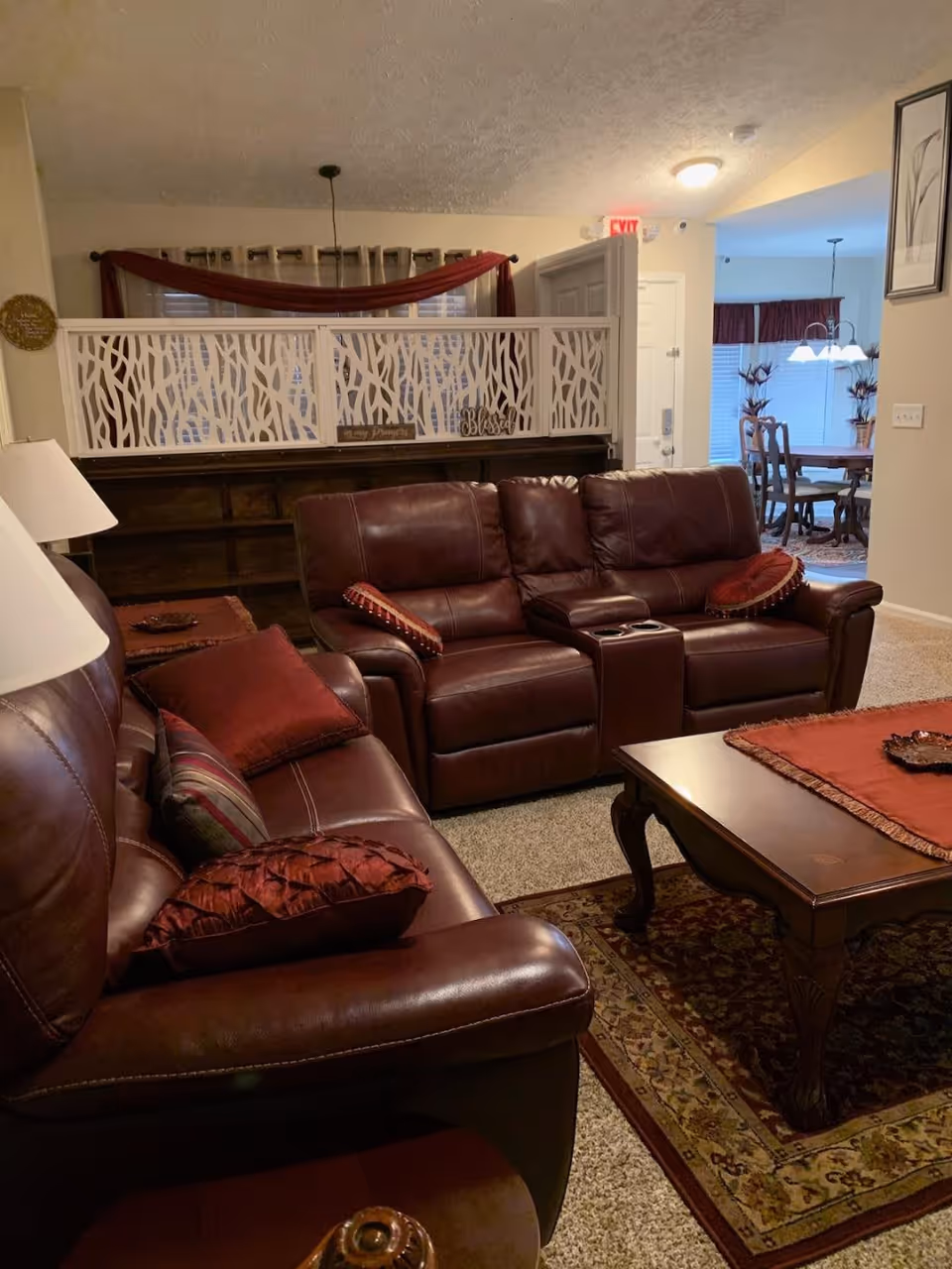 A cozy living room with dark brown leather sofas adorned with red and patterned pillows. A wooden coffee table with a red runner sits on a patterned rug. Behind the sofas is a decorative white room divider with a nature-inspired cutout design. In the background, a dining area with a wooden table and chairs is visible, along with a hanging light fixture and window treatments with red curtains.