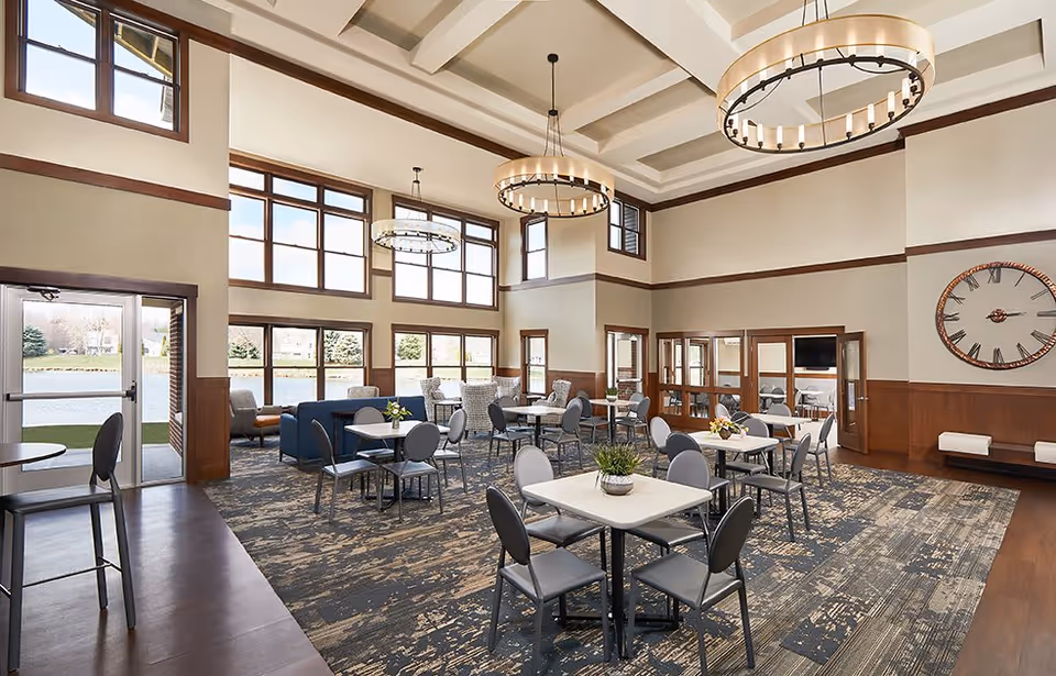 A spacious and well-lit common area in a senior living facility with multiple tables and chairs arranged for dining or socializing. The room features large windows allowing natural light to fill the space, modern circular chandeliers hanging from a high coffered ceiling, and a large wall clock. There are also some lounge chairs and a blue sofa near the windows, with a door leading outside to a grassy area.