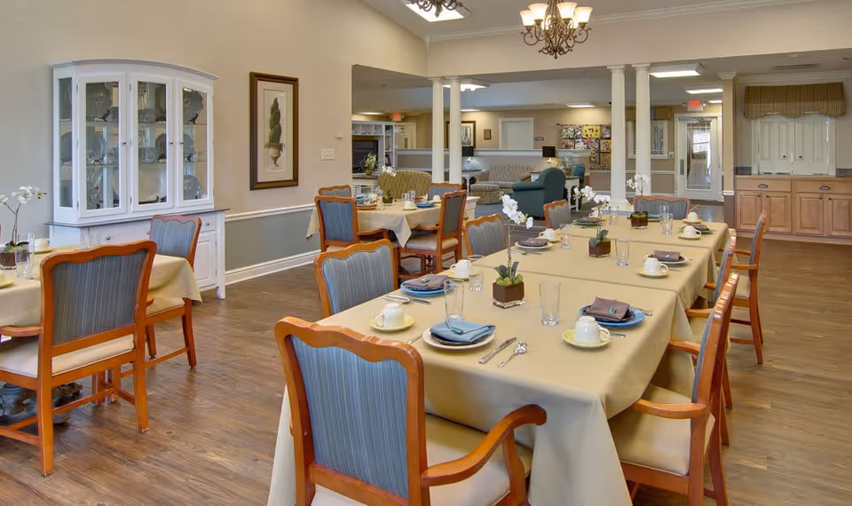 A dining room in a senior living facility with tables covered in beige tablecloths, set with plates, cups, glasses, and silverware. Wooden chairs with blue cushions surround the tables. The room has wooden flooring, a white cabinet with glass doors displaying decorative items, framed artwork on the wall, and a chandelier hanging from the ceiling. In the background, there is a seating area with sofas and a bulletin board.