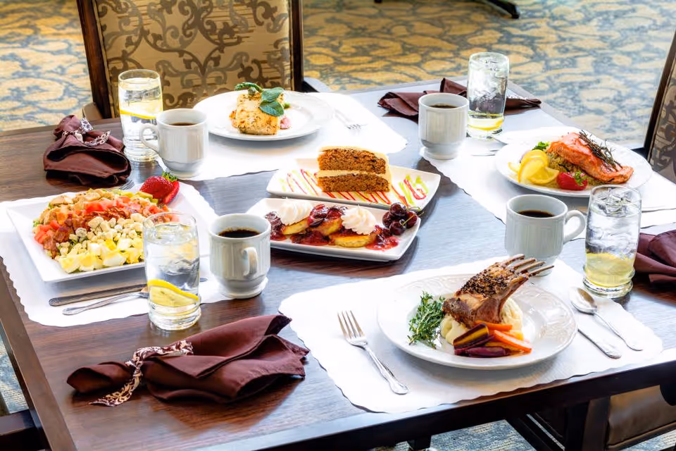 A dining table set with various plated meals including a salad, grilled salmon with lemon, a rack of lamb with mashed potatoes and vegetables, a dessert plate with cake and fruit-topped pastries, along with cups of coffee and glasses of water with lemon. The table has brown napkins and is in a room with patterned carpet and upholstered chairs.