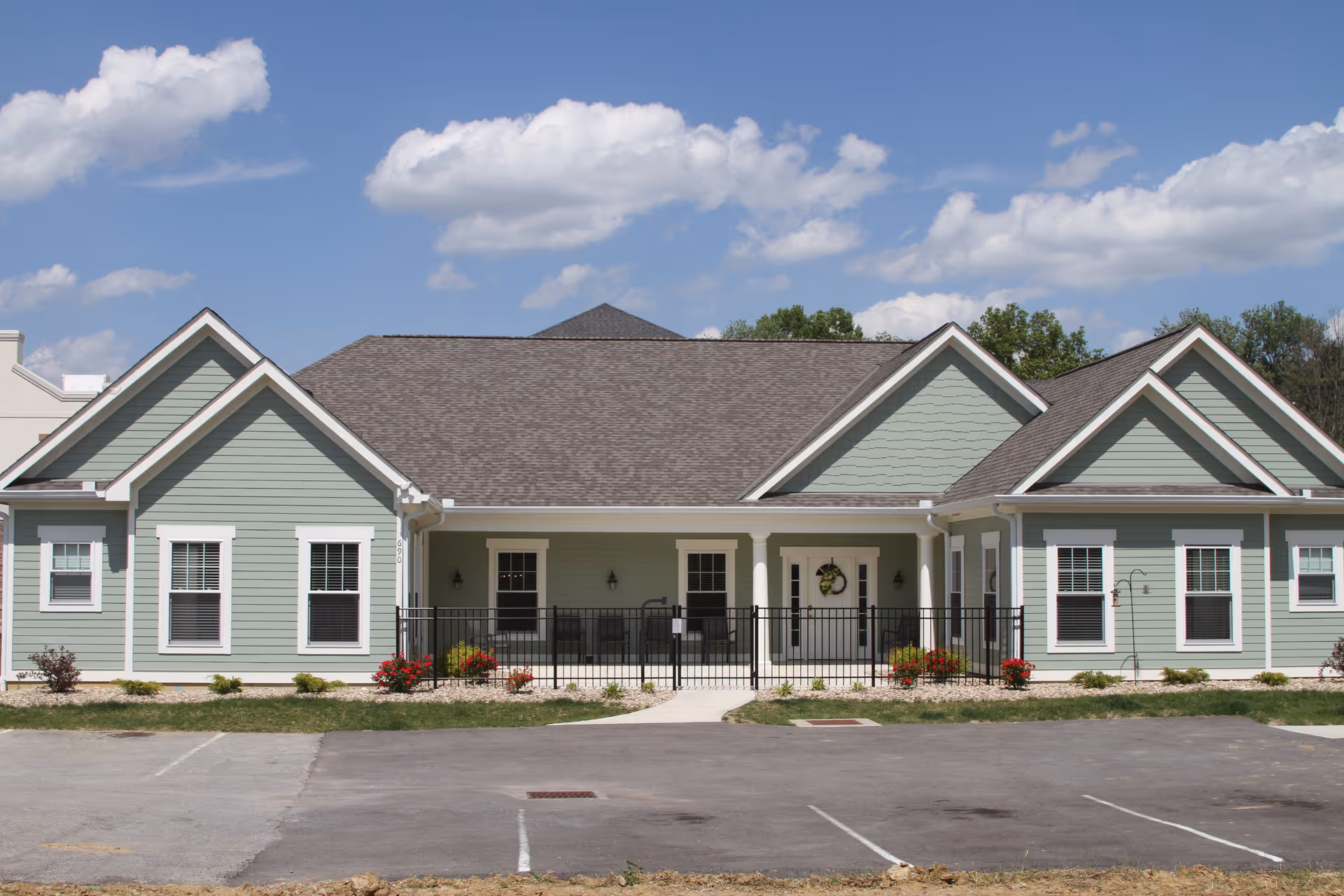 Front exterior view of a single-story building with light green siding, white trim, and a gray shingled roof under a blue sky with scattered clouds. The building has multiple windows, a central entrance with a small porch, and a black metal fence enclosing the porch area. There are small shrubs and flowers planted along the front of the building, and an empty parking lot in the foreground.