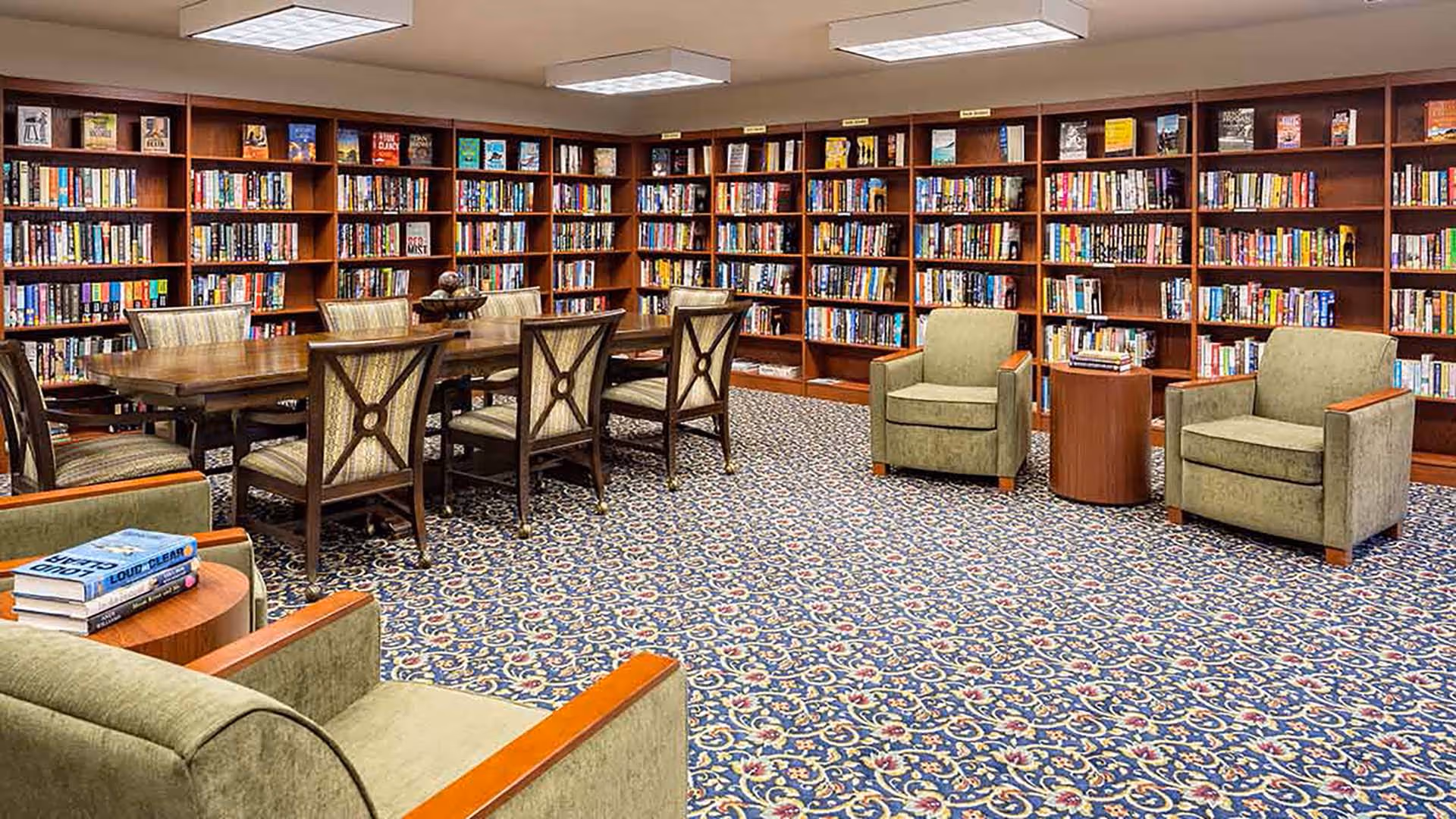 A cozy library room with wooden bookshelves filled with books lining the walls. In the center, there is a long wooden table surrounded by cushioned chairs. There are also several comfortable armchairs with wooden armrests and small round tables beside them. The room has a patterned carpet and bright overhead lighting.