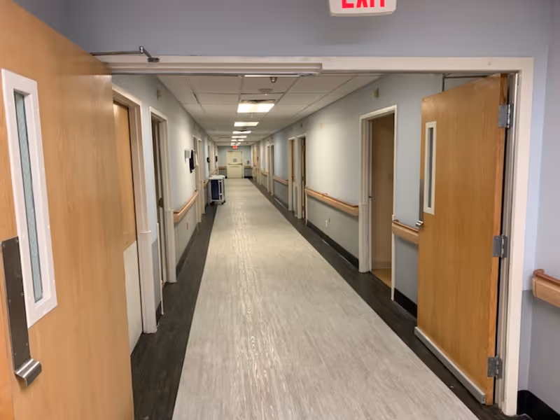 A long, clean hallway in a senior living or rehabilitation facility with light-colored walls, wood handrails on both sides, and several open wooden doors leading to rooms. The floor has a light center strip with darker edges, and fluorescent lights illuminate the ceiling. An exit sign is visible at the far end of the hallway.