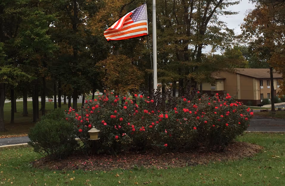 An American flag on a pole above a bed of red rose bushes in front of a two-story beige building and trees.
