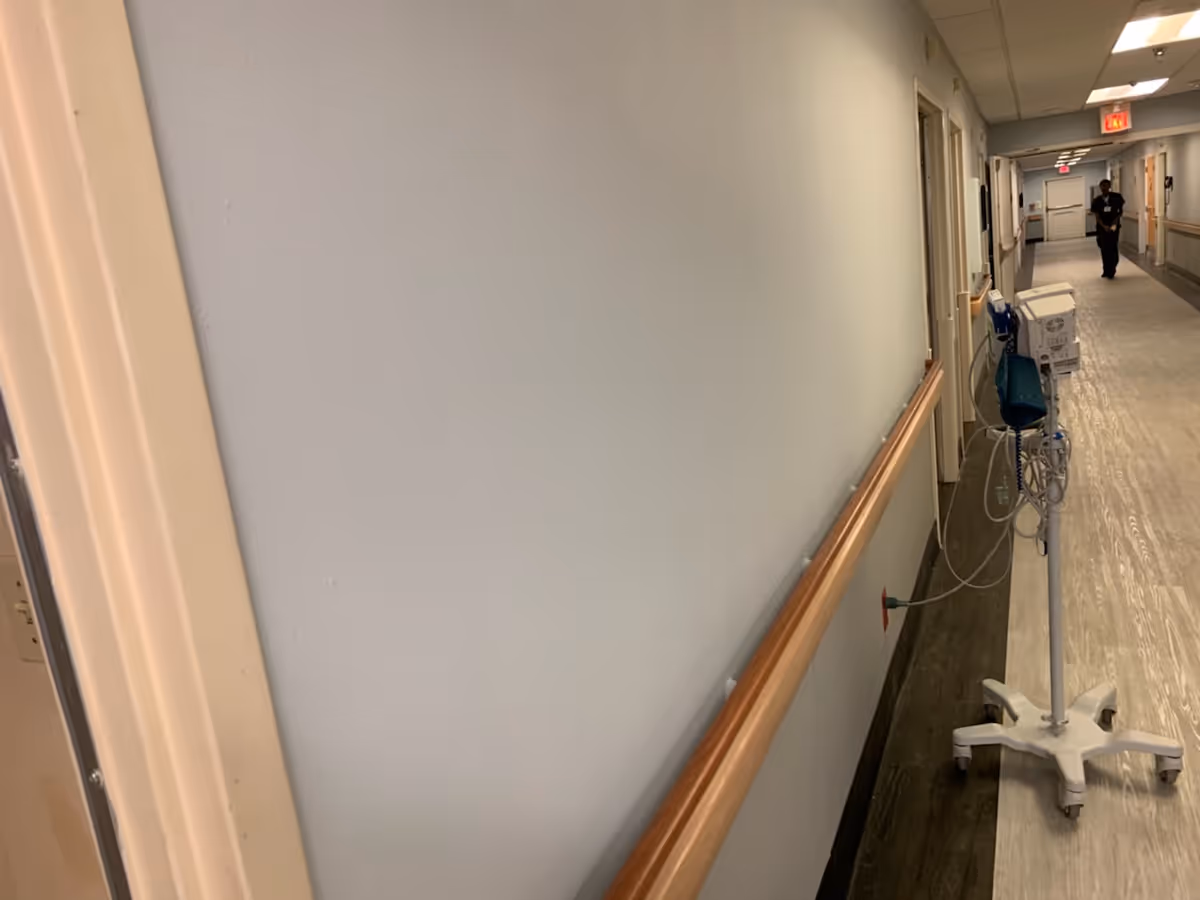 A long, empty hallway in a senior living facility with light-colored walls and a wooden handrail on the right side. Medical equipment on a wheeled stand is positioned along the hallway. A person is seen walking in the distance near an exit sign.