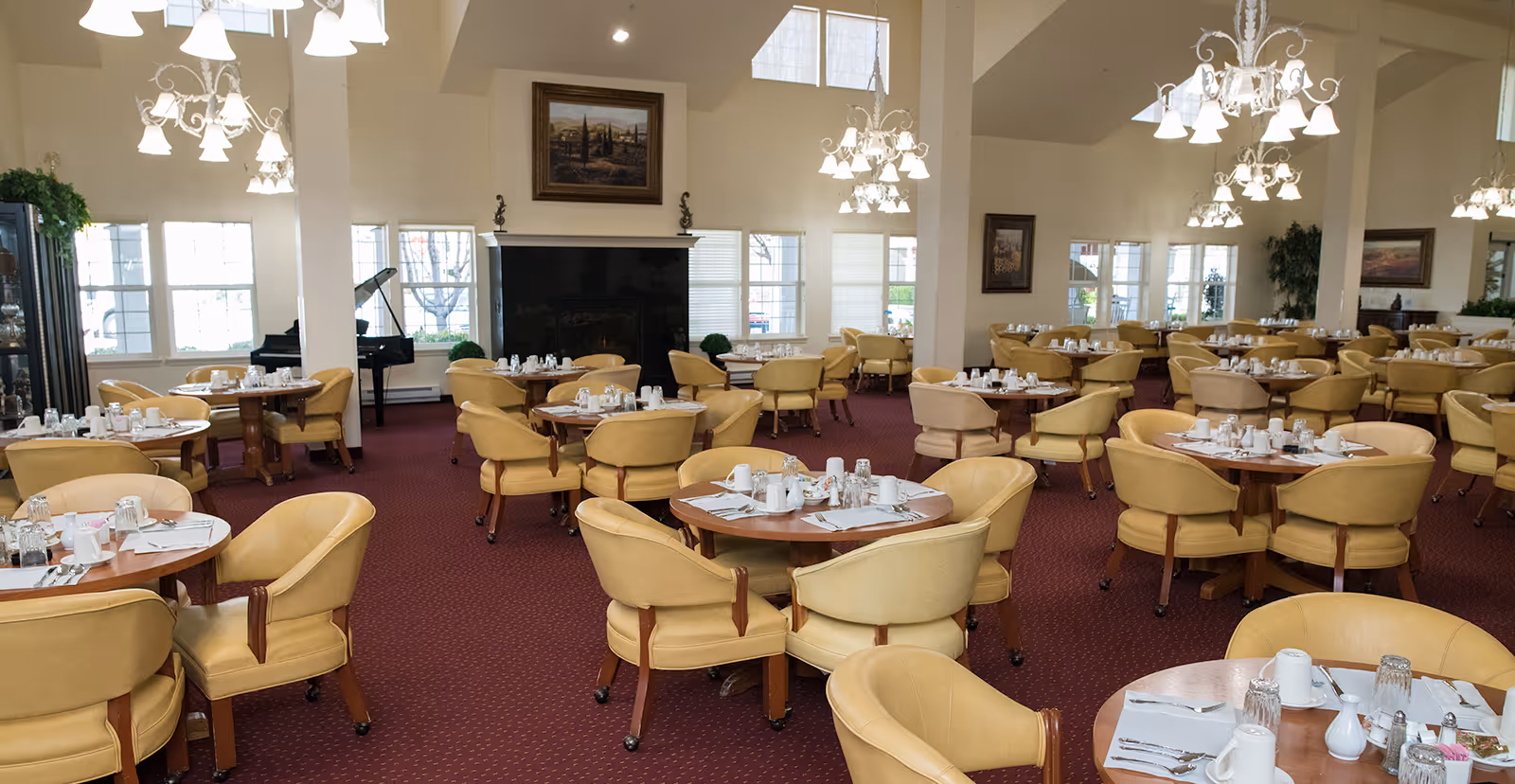 A spacious dining room with multiple round wooden tables surrounded by beige cushioned chairs. Each table is set with white cups, glasses, napkins, and silverware. The room has a red carpet, large windows letting in natural light, chandeliers hanging from the ceiling, and a black fireplace with a framed painting above it. There is also a black grand piano near the windows.