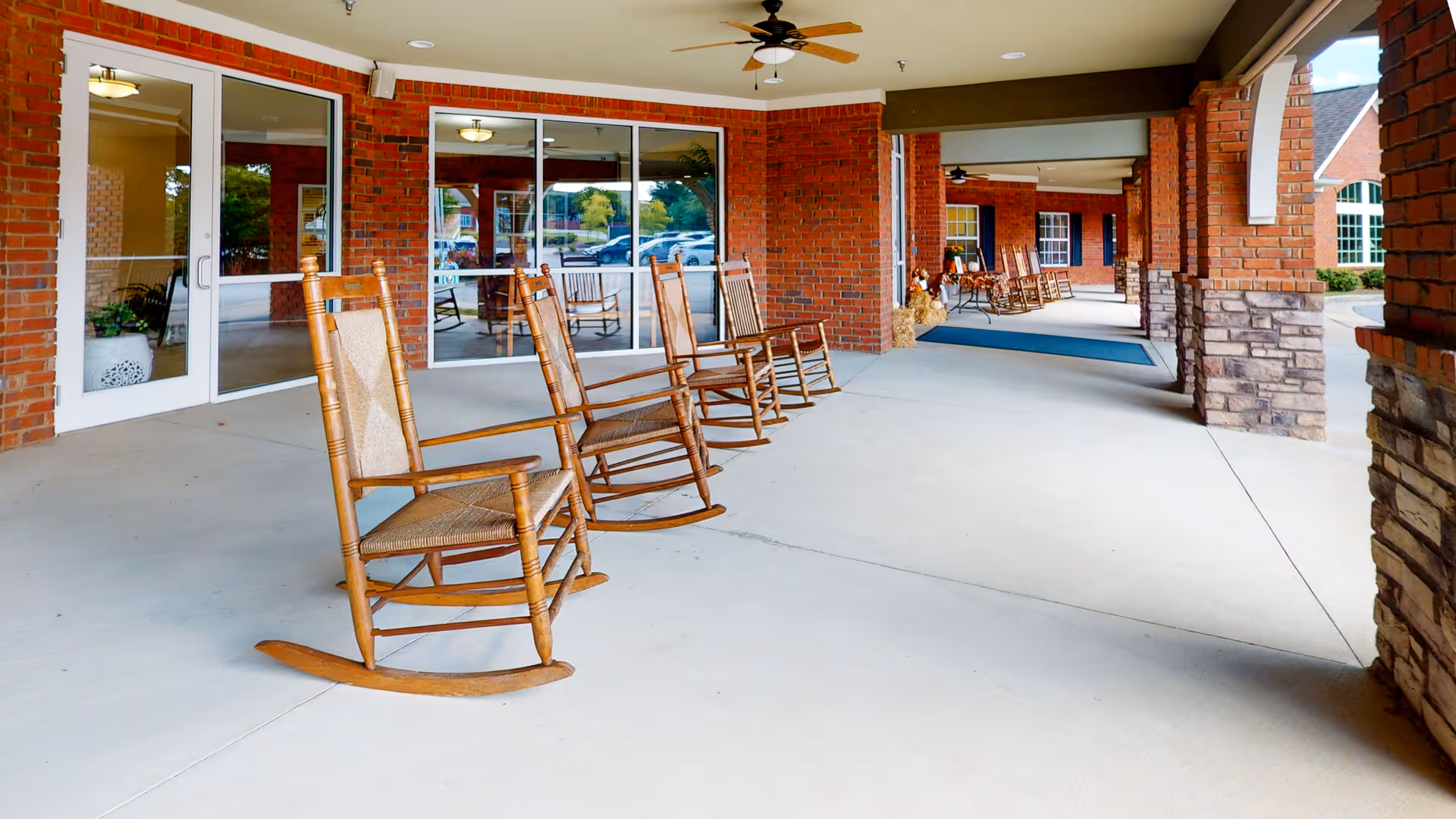 Covered outdoor patio area at Gaines Park Senior Living with several wooden rocking chairs arranged along a brick wall with large windows and glass doors. The patio has a concrete floor and ceiling fans, with a view of parked cars and greenery outside.