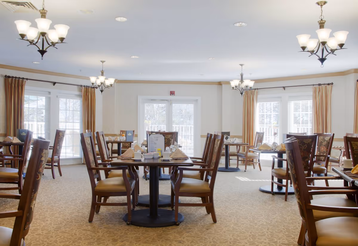 Bright dining room with multiple tables set for meals, wooden chairs, chandeliers, and large windows with curtains.