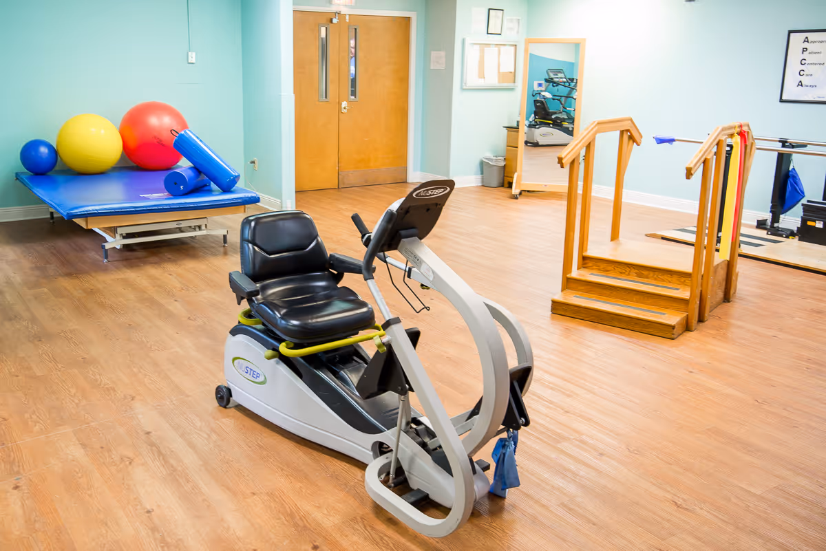 A physical therapy or rehabilitation room with exercise equipment including a recumbent exercise bike, wooden stairs with handrails for step practice, a blue exercise mat with large colorful exercise balls, and a large mirror on the wall. The room has light blue walls and wooden flooring.