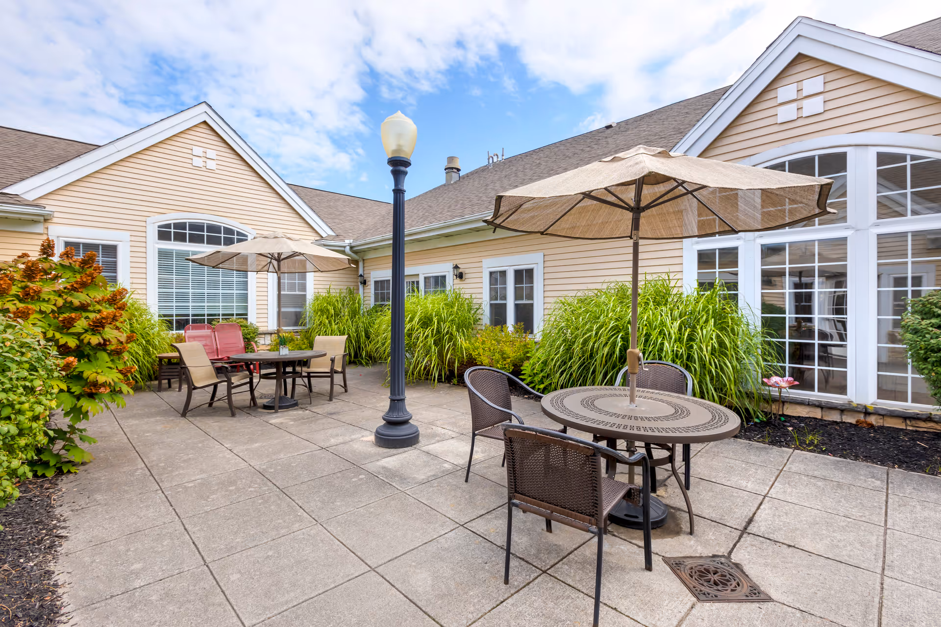 Outdoor patio area at Brookdale Fayetteville with two round tables, each shaded by a large beige umbrella. The patio is paved with concrete tiles and surrounded by green shrubs and plants. The building exterior is light beige with white trim and large windows. A black lamppost stands in the middle of the patio.