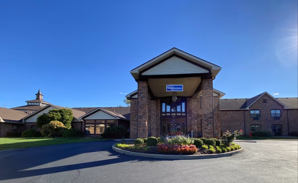Front exterior view of a brick building with a covered entrance and a 'Welcome' sign hanging above the doorway. There is a circular flower bed with various plants and flowers in front of the entrance. The sky is clear and blue.