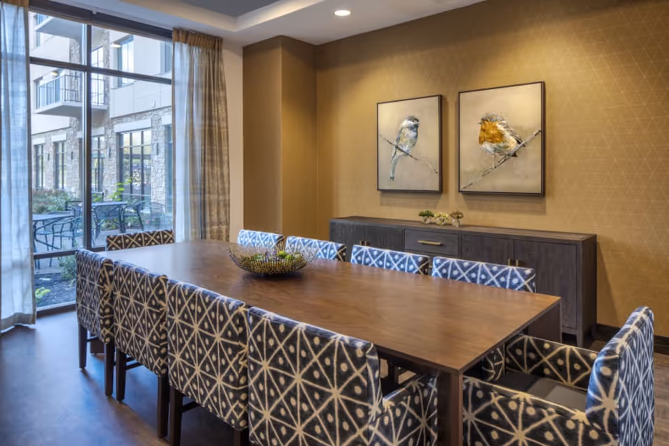 Well-appointed dining room with a long wooden table surrounded by patterned upholstered chairs, a sideboard with bird paintings, and a large window overlooking an outdoor patio.