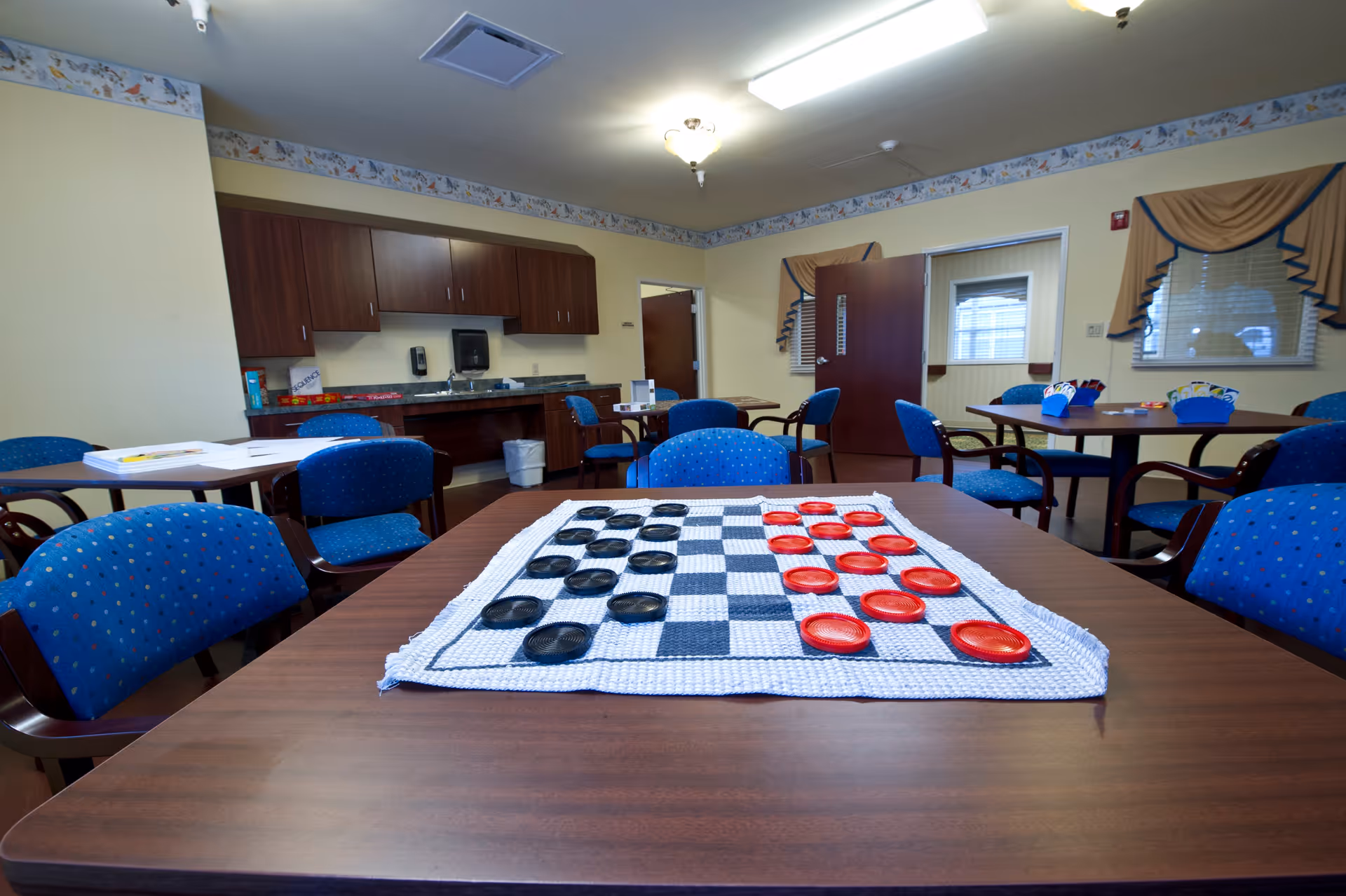 A common activity room with several tables and blue cushioned chairs. On the nearest table, there is a large checkers board with red and black pieces set up. The room has light yellow walls, wooden cabinets, a sink, and windows with beige curtains. The door is open, showing another room beyond.