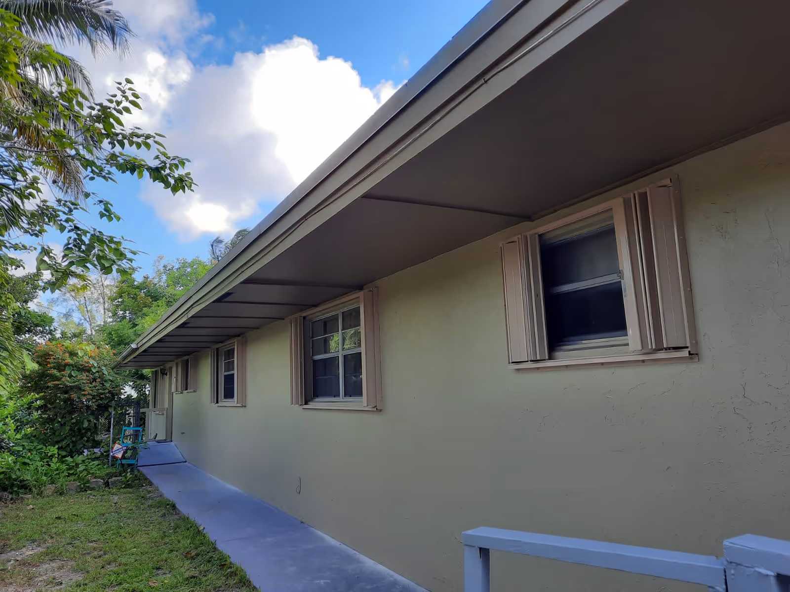 Exterior side view of a single-story building with beige walls and several windows with shutters. There is a concrete walkway along the building and greenery including trees and bushes nearby under a partly cloudy sky.