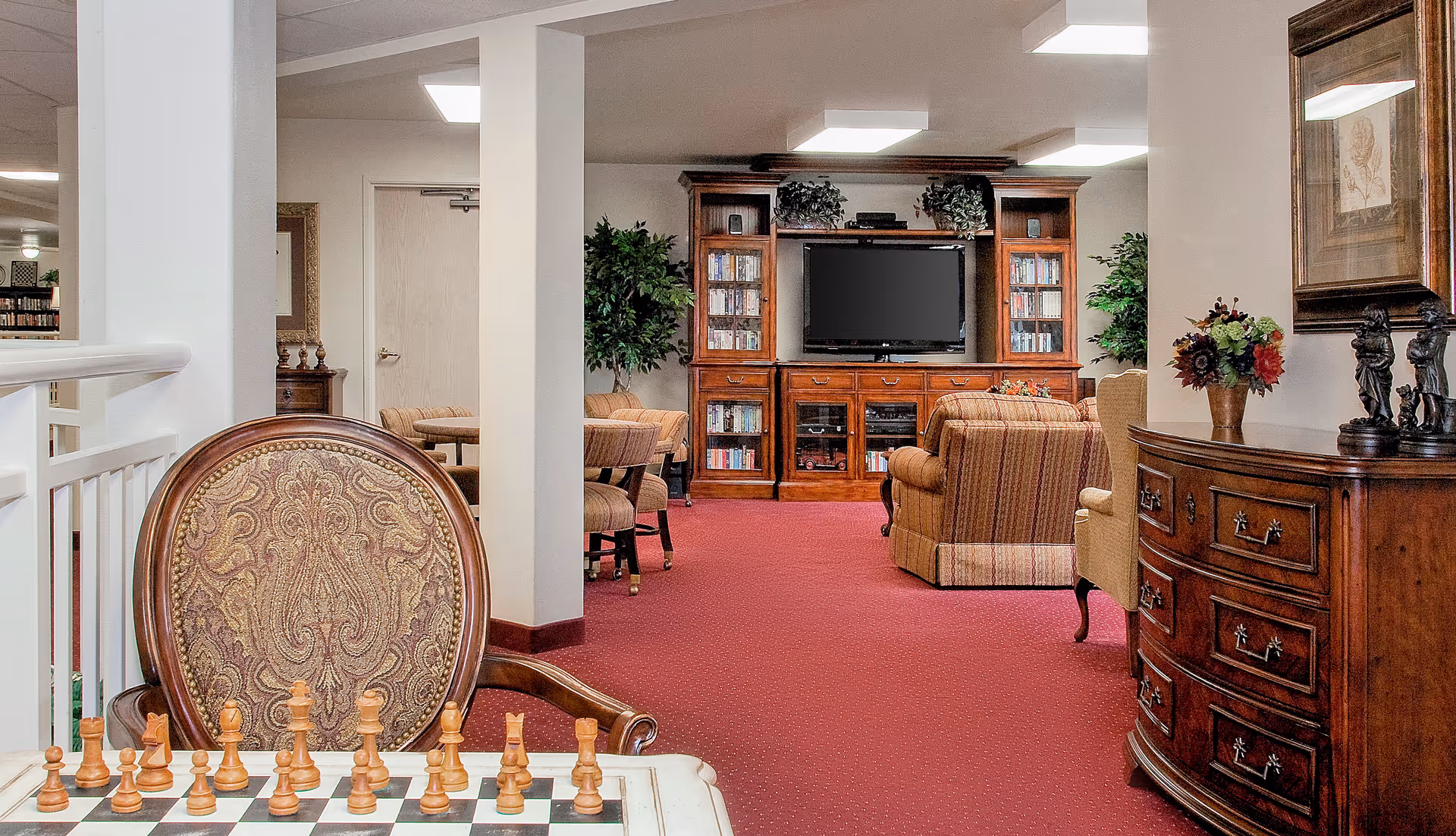 Cozy common living area with a chessboard in the foreground, upholstered chairs, and a wooden entertainment center with a TV.