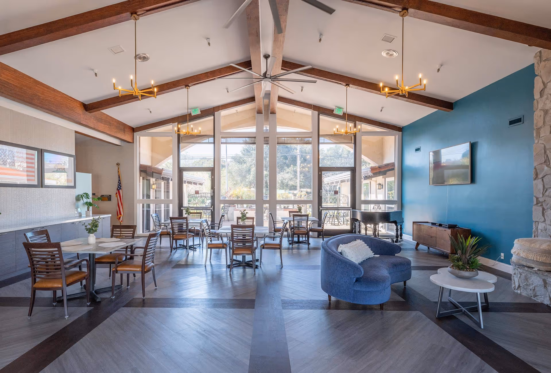 A bright communal lounge and dining area with multiple tables and chairs, a blue circular sofa, TV, piano, and large floor-to-ceiling windows under exposed wooden beams.