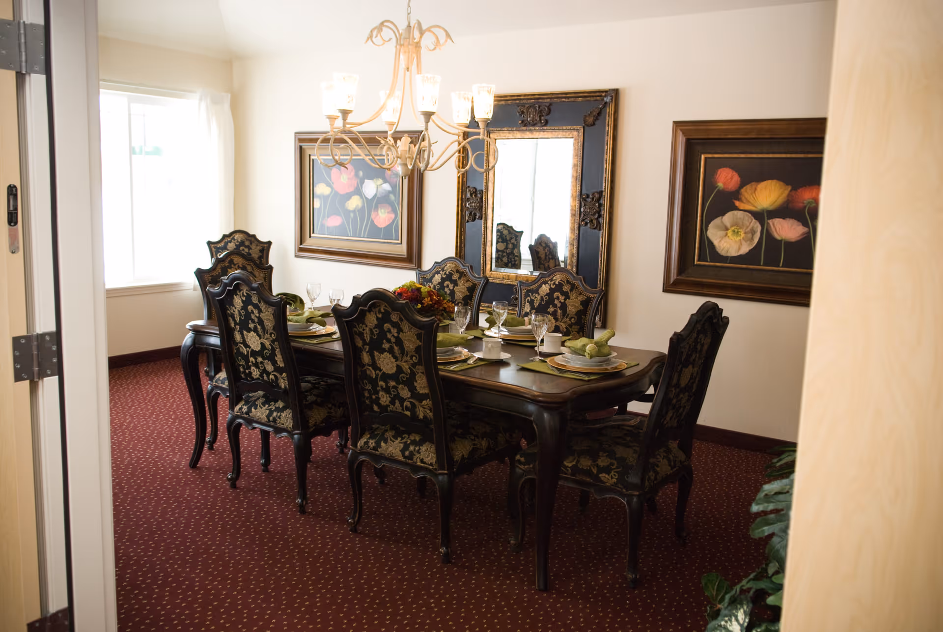 A formal dining room with a dark wooden table set for six people. The chairs have ornate black and gold upholstery. The room has a red carpet with a subtle pattern, two framed floral paintings on the walls, and a large decorative mirror. A chandelier with multiple lights hangs above the table, and a window with white curtains lets in natural light.
