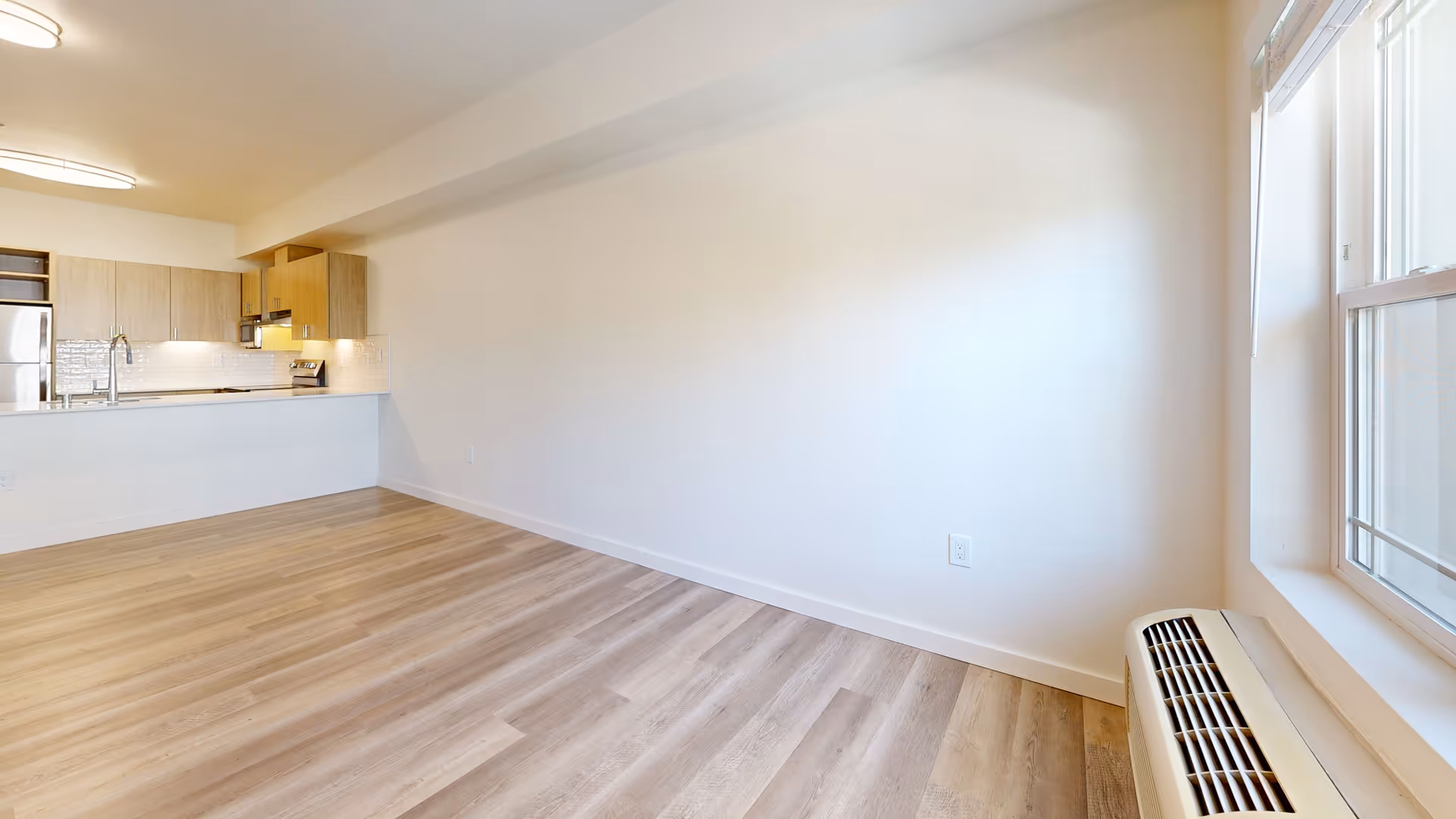 Empty room with light wood flooring, white walls, and a large window on the right side. The room opens into a kitchen area with light wood cabinets, a stainless steel refrigerator, a stove, and a sink with a modern faucet. The space is well-lit with ceiling lights and natural light from the window.