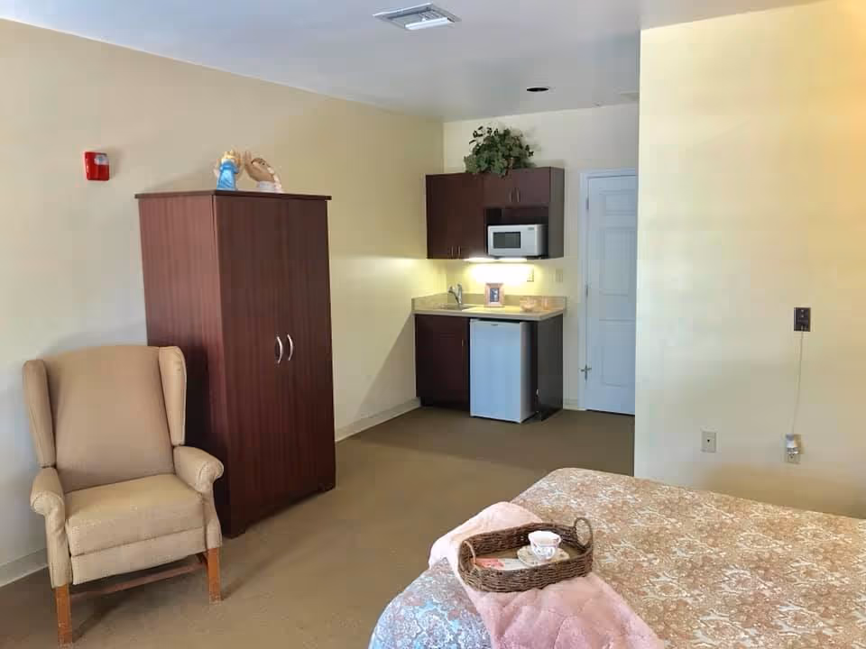 A cozy room with a beige armchair, a dark wooden wardrobe, a small kitchenette with a microwave, mini fridge, and cabinets, and a bed with a patterned bedspread and a tray holding a teacup and saucer.