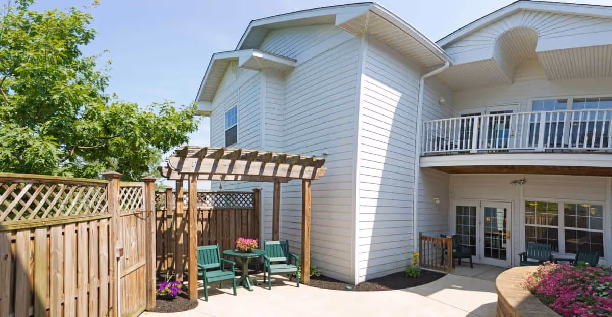 Outdoor patio area at Newhaven Court at Lindwood featuring a wooden pergola with a small table and green chairs underneath, surrounded by a wooden fence and greenery. The building exterior is white with multiple windows and a balcony above the patio. There are flower beds with pink flowers near the patio.