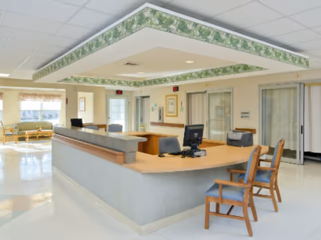 Reception area in a senior living facility with a large desk, computer, chairs, and a seating area with windows in the background. The ceiling has a decorative border with a floral pattern.