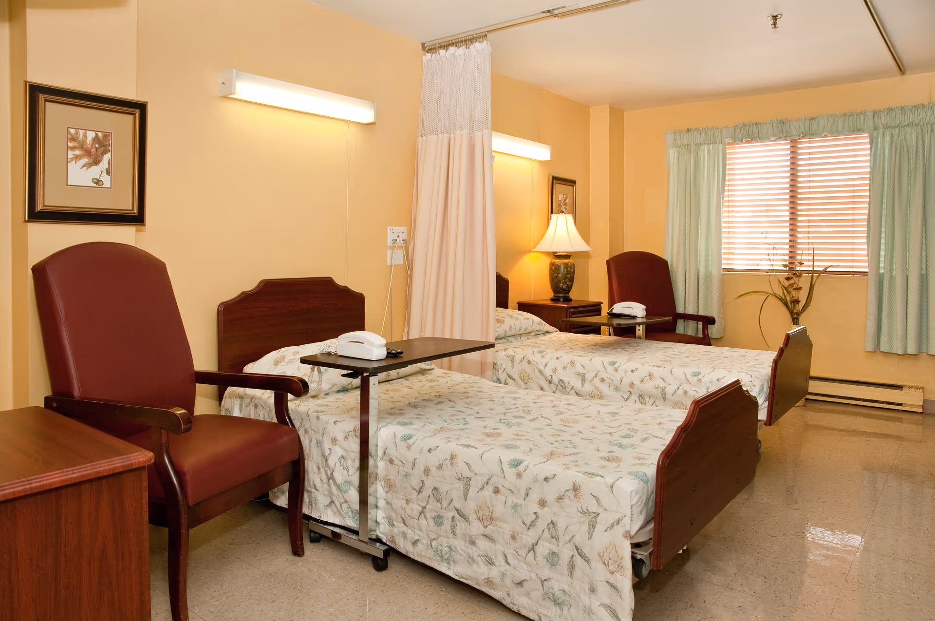 A room with two hospital-style beds covered with floral bedspreads, each with an overbed table holding a white telephone. There are two maroon armchairs, a wooden nightstand with a lamp, a framed picture on the wall, and a window with green curtains and blinds. A privacy curtain is partially drawn between the beds.