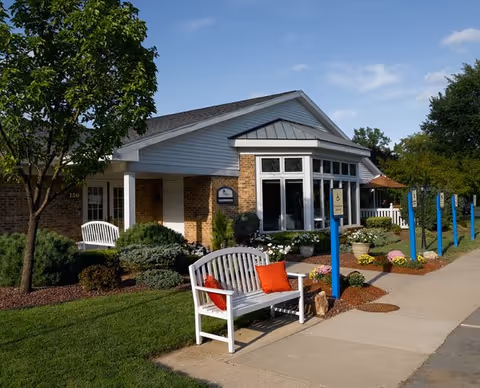 Front exterior of a brick senior living community building with a white bench, landscaped flowerbeds, and a sidewalk.