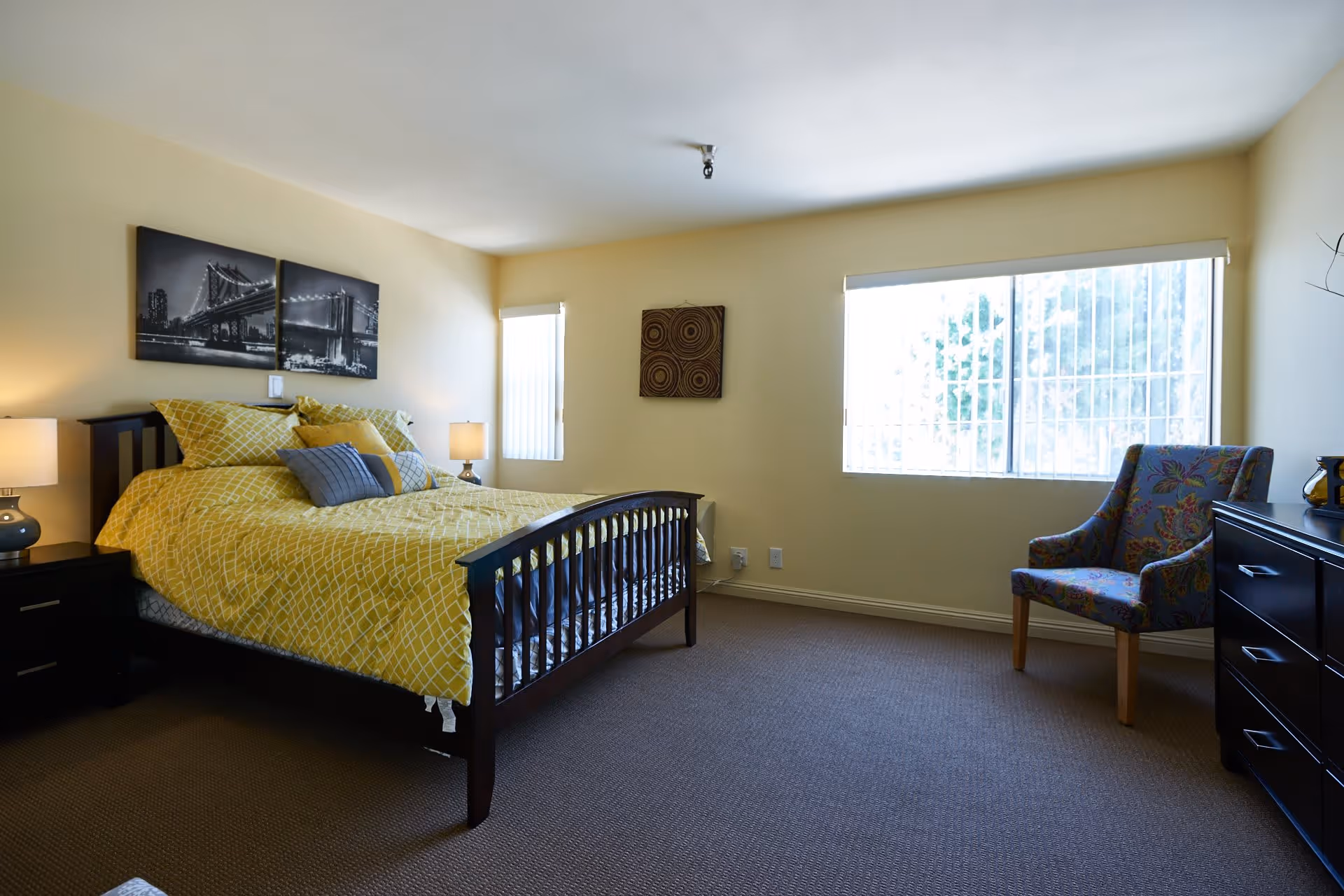 A bedroom with a dark wooden bed frame and yellow patterned bedding. There are two bedside tables with lamps, a colorful upholstered armchair, a dark wooden dresser, and two windows with vertical blinds letting in natural light. The walls are painted light yellow and decorated with black and white cityscape artwork and a circular patterned piece.