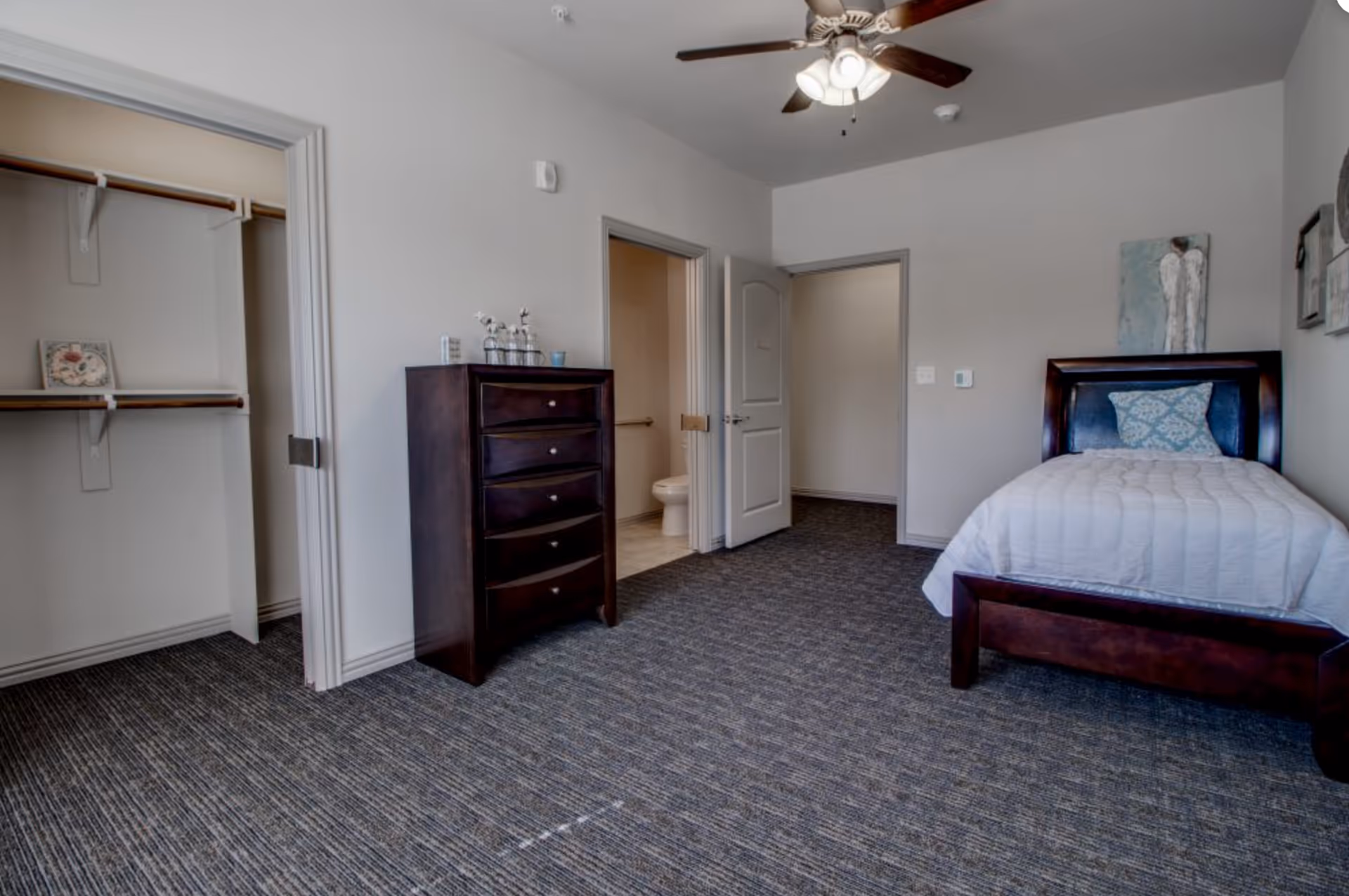 A furnished bedroom with a single bed, dark wood dresser, open closet, and a doorway leading to a bathroom.