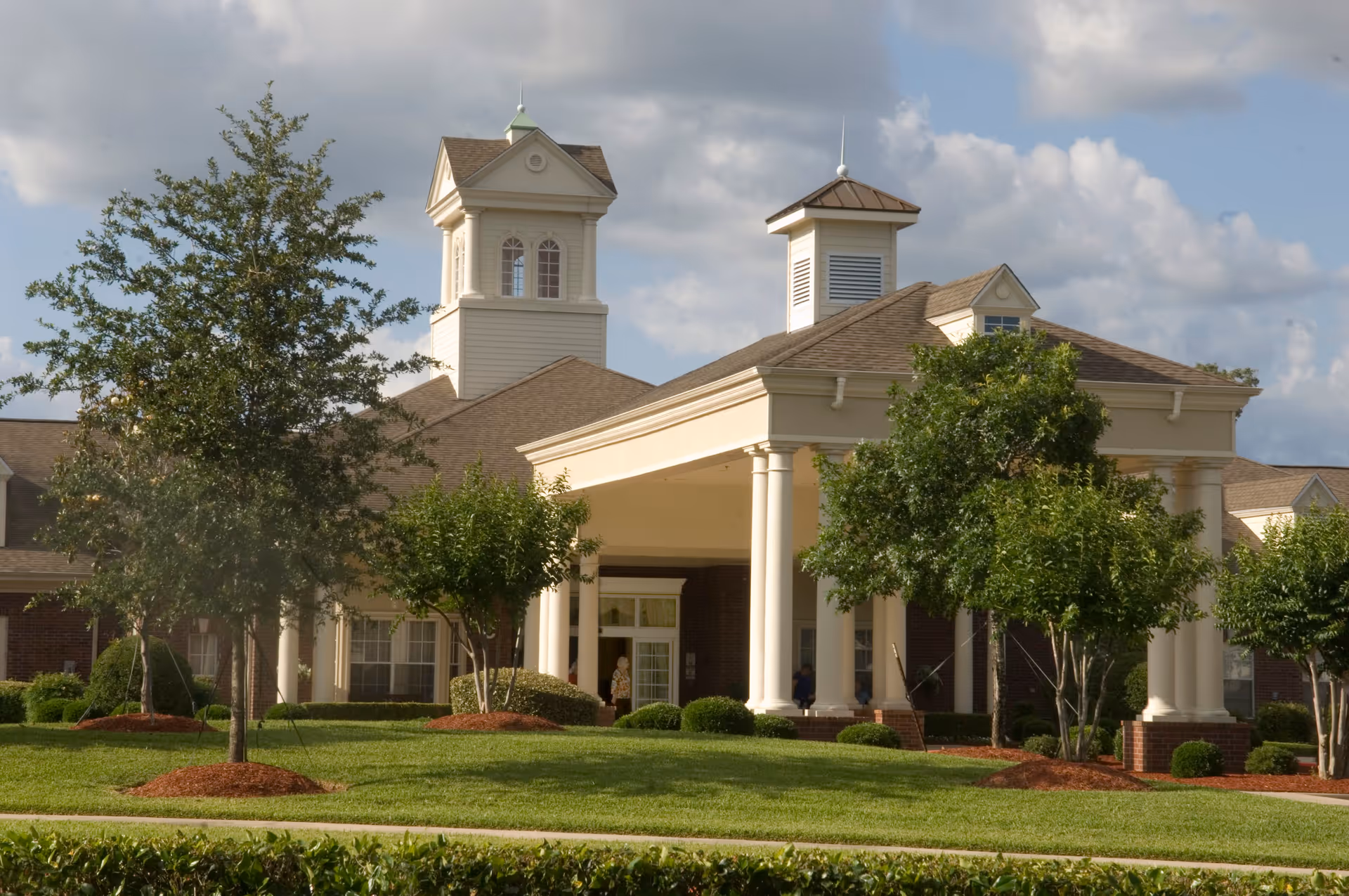 Front exterior view of a senior living facility building with a covered entrance supported by white columns, manicured green lawn, several trees, and a partly cloudy sky in the background.