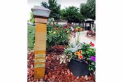 Outdoor garden area with a wooden post, various colorful flowers including petunias and other plants, red mulch covering the ground, and trees and a building visible in the background under a clear sky.