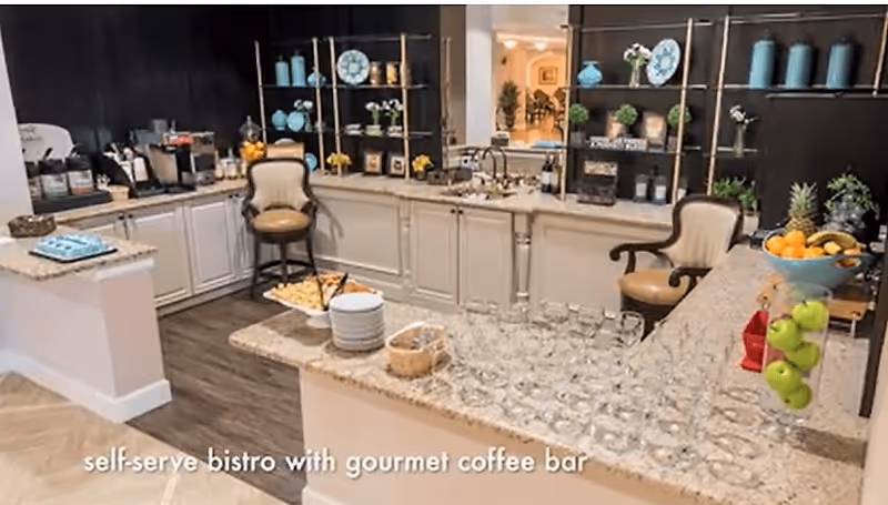 Interior view of a self-serve bistro with a gourmet coffee bar featuring granite countertops, glassware, plates, snacks, and decorative shelves with vases and plates. Two cushioned chairs are placed near the counter.
