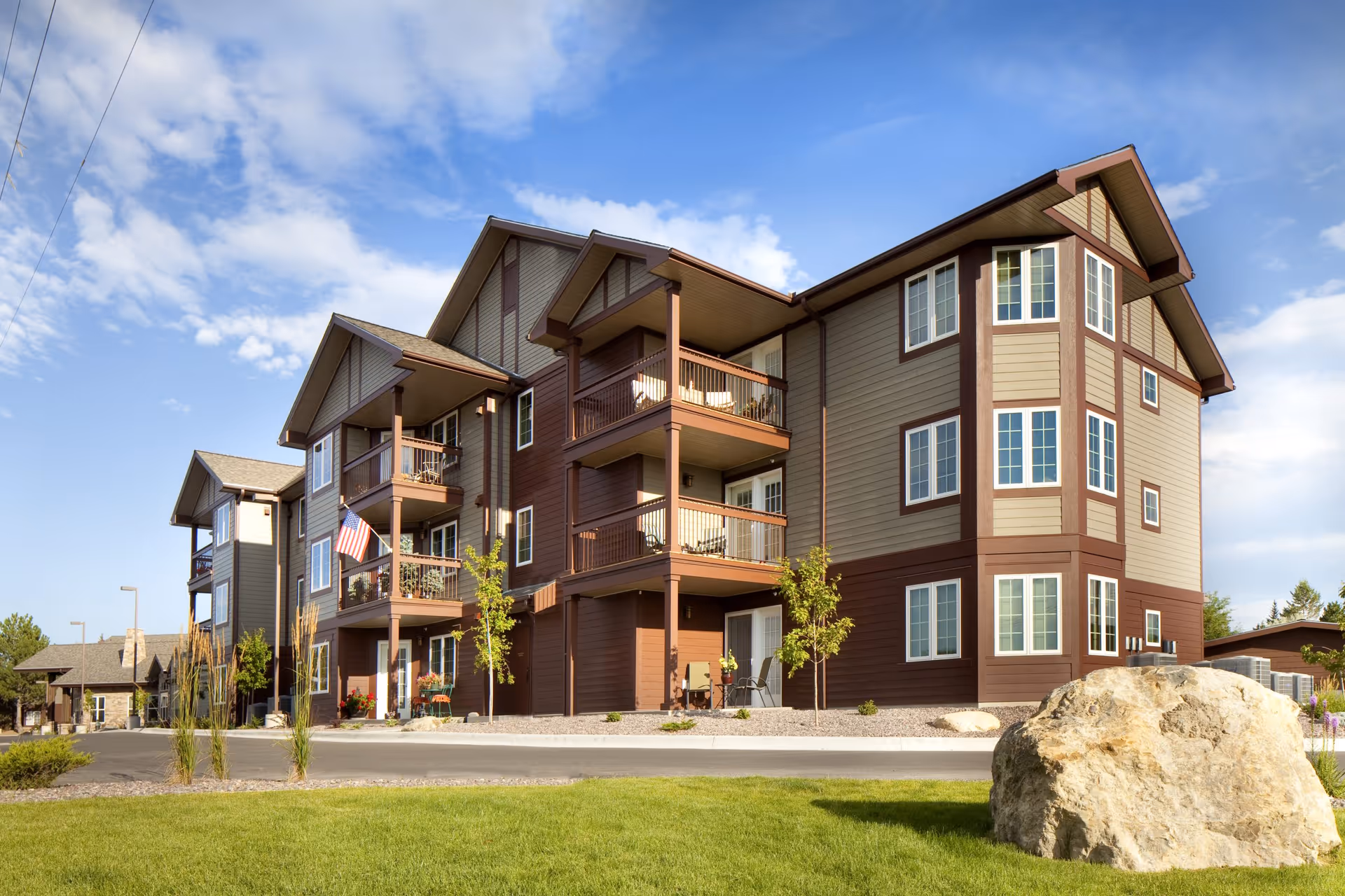 Exterior view of a three-story residential building with balconies, an American flag, a manicured lawn and a large rock under a blue sky.