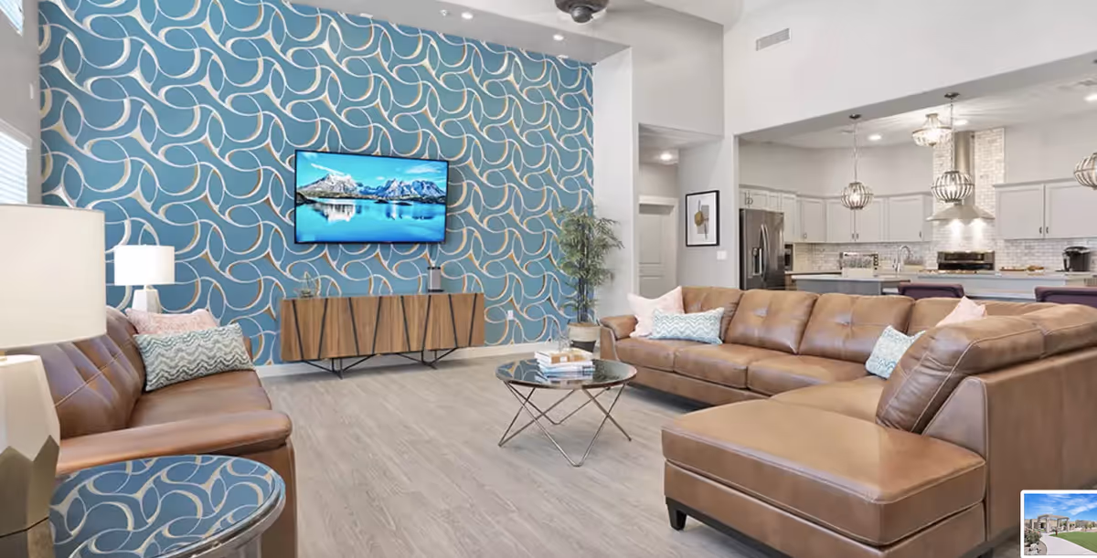 A modern living room with brown leather sofas, a round glass coffee table, and a wooden TV stand against a blue patterned accent wall. The room opens into a kitchen area with white cabinets, stainless steel appliances, and pendant lights.