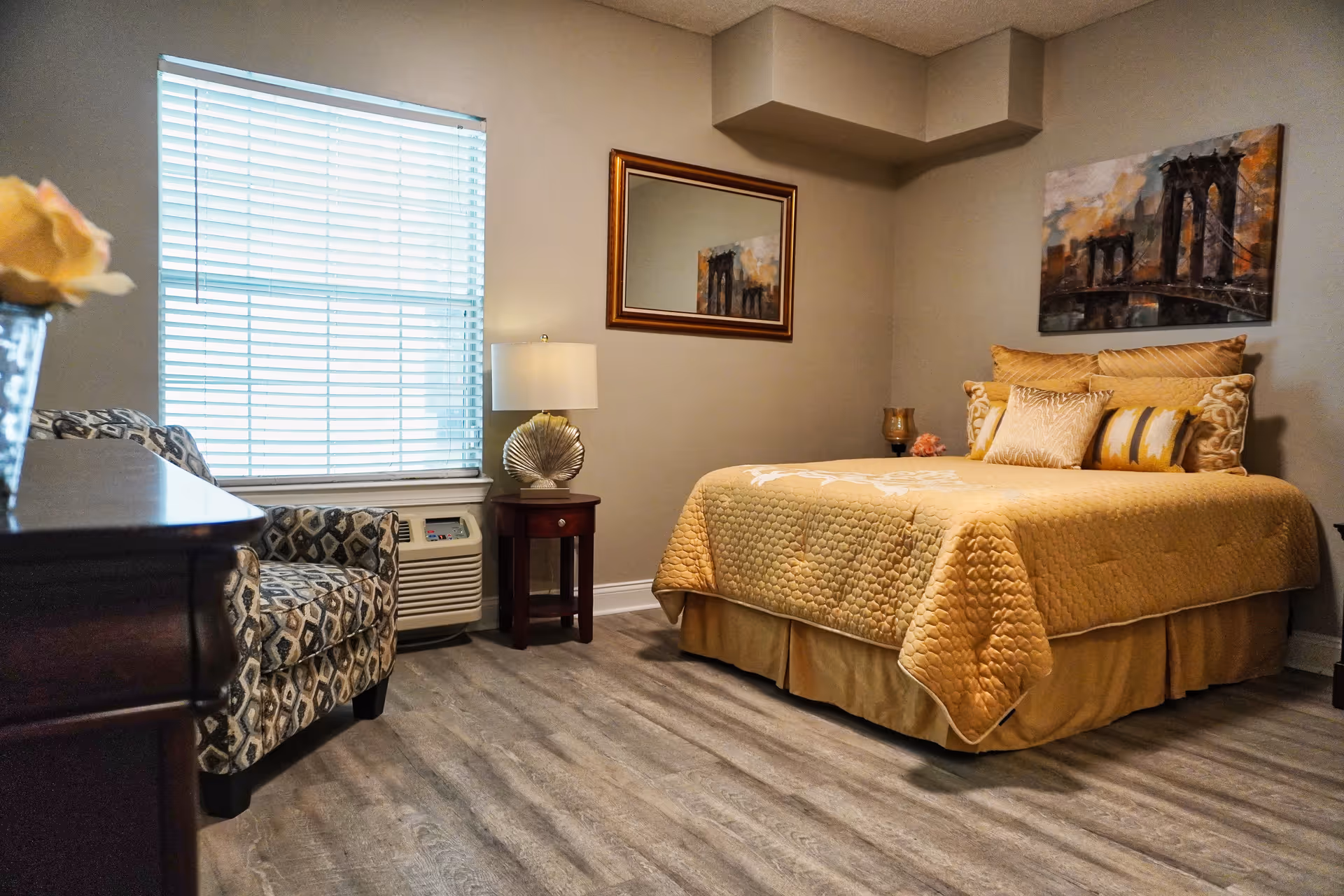 A cozy bedroom with a bed covered in a golden quilt and multiple pillows. There is a patterned armchair near a window with blinds, a small wooden side table with a decorative lamp, and a framed mirror on the wall reflecting part of a painting of a bridge.