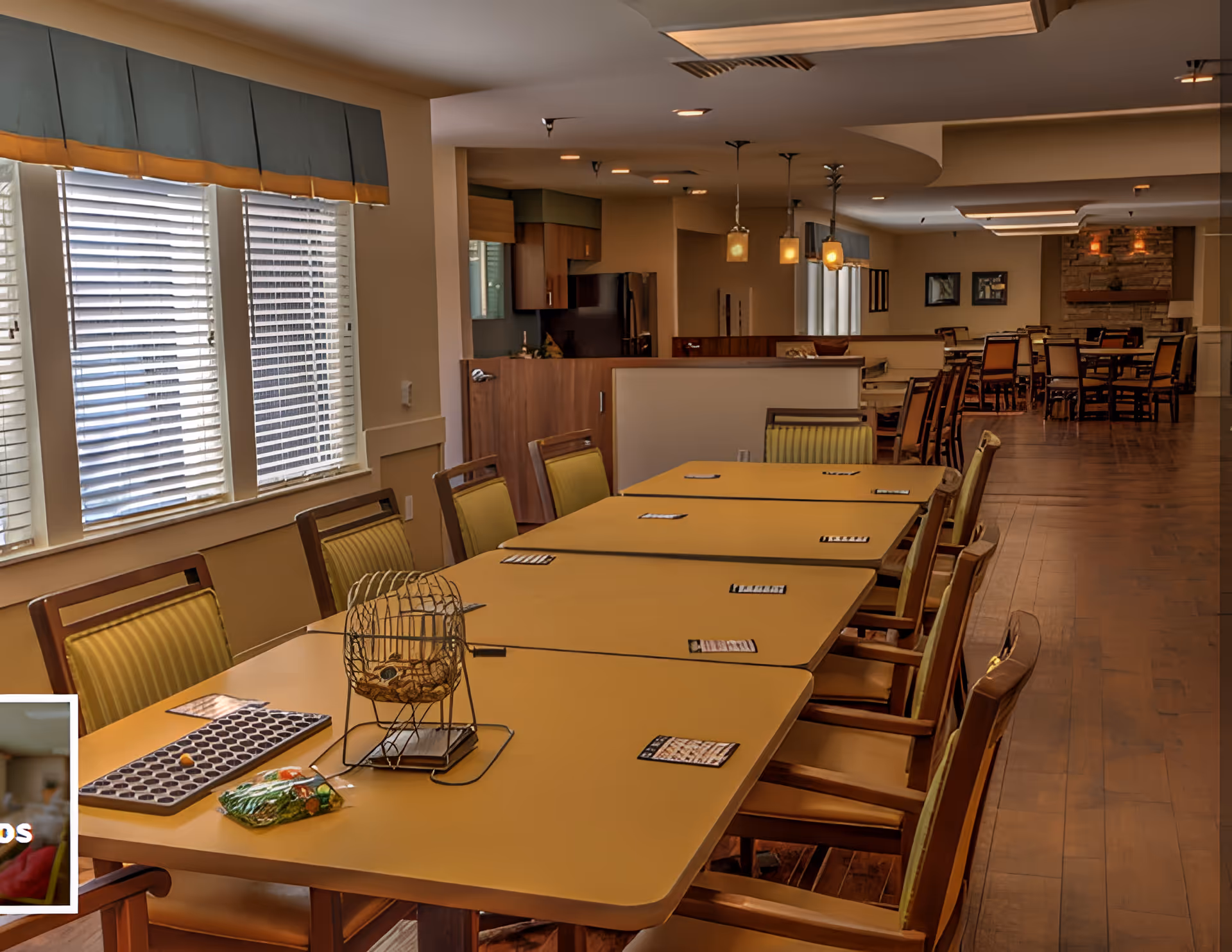 Spacious communal dining/activity room with long tables and chairs set up for games including a bingo cage and cards, windows and pendant lights.