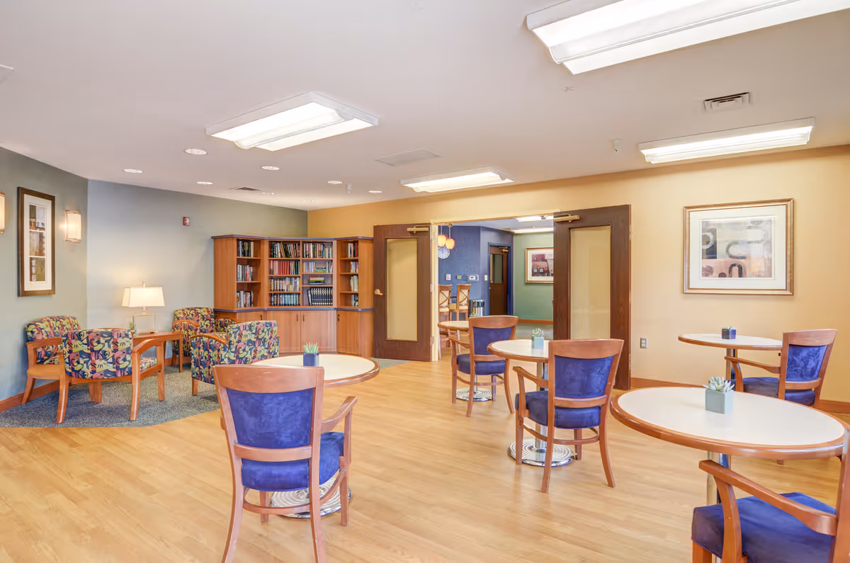 A well-lit common area in a senior living facility with several round tables and wooden chairs with blue cushions. In the background, there is a cozy reading nook with colorful armchairs, a wooden bookshelf filled with books, and a table lamp. The walls are painted in soft tones with framed artwork, and double wooden doors lead to another room.