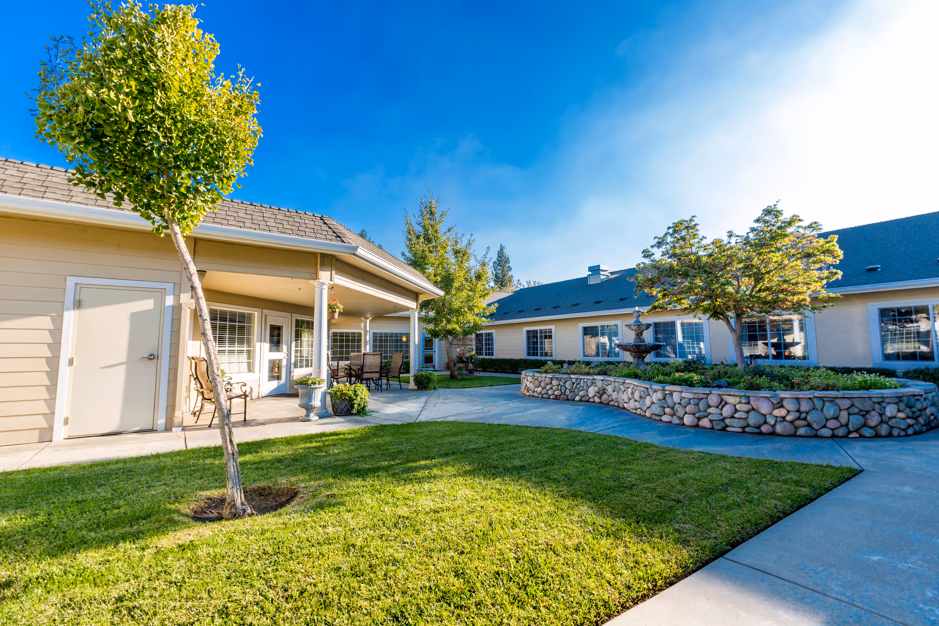 Sunny courtyard with a grassy lawn, stone planter with a fountain, patio seating and surrounding single-story buildings.