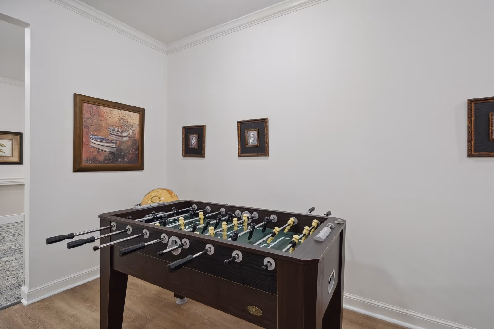 A foosball table sits in a small interior room with white walls and framed artwork.