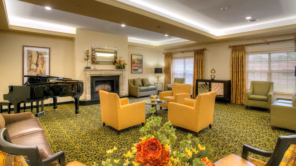 A spacious living room with patterned carpet, four yellow armchairs arranged around a small round glass table, a grand piano on the left, a fireplace with a decorative mirror above it, and several couches and armchairs near windows with yellow curtains. There are framed paintings on the walls and a floral arrangement in the foreground.
