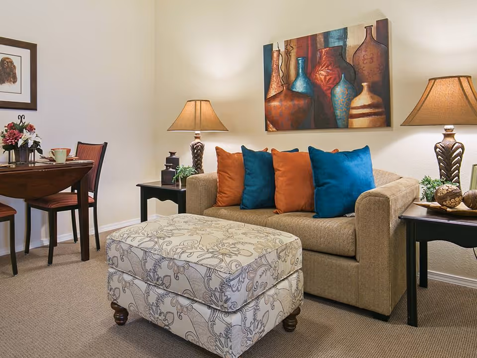 Cozy living room with a beige sofa, colorful throw pillows, a patterned ottoman, side tables and lamps beneath wall art.