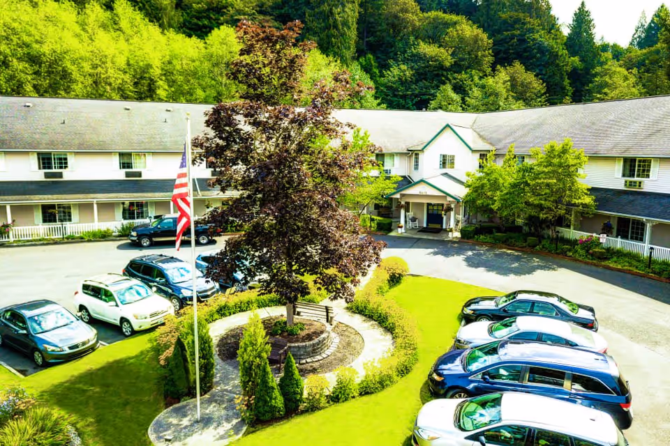 Exterior view of Cascade Valley Senior Living facility showing a two-story building with a gray roof surrounded by lush green trees. In front of the building is a circular driveway with several parked cars and a landscaped area with a flagpole flying the American flag, a large tree, and a bench.