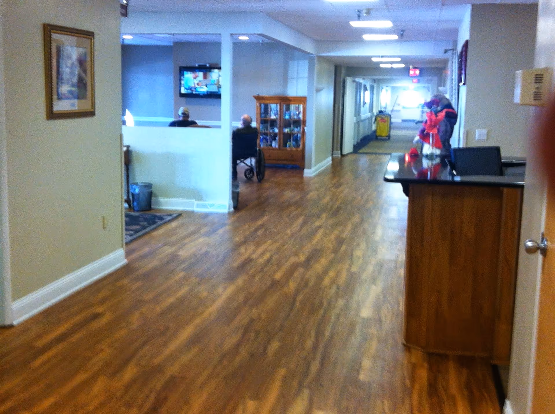 Interior hallway of a senior living facility with wood flooring, a reception desk on the right, a display cabinet in the middle, and two people sitting in wheelchairs near a partitioned area with a television mounted on the wall.