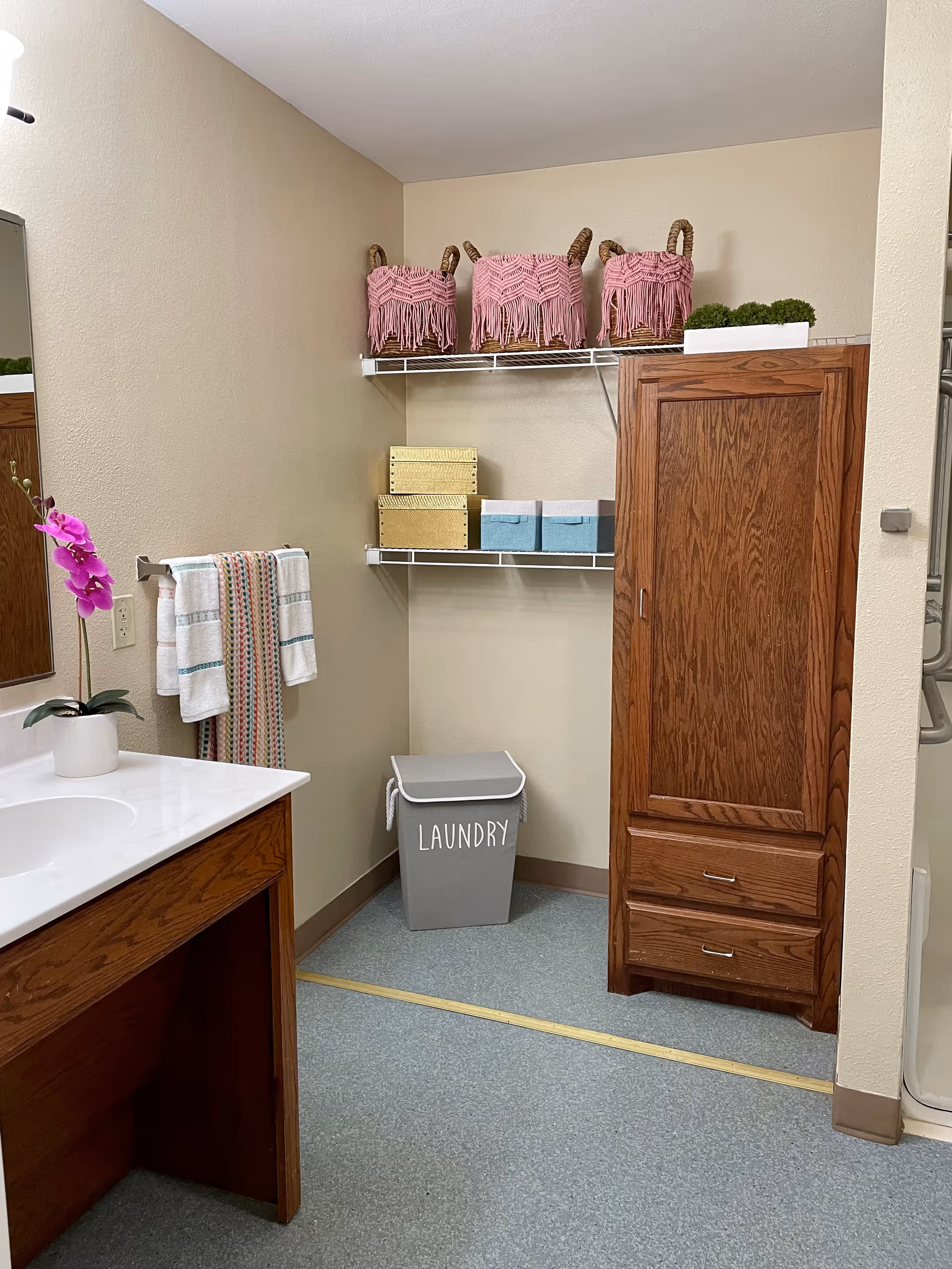 A tidy assisted-living bathroom with a sink and vanity, wall shelves holding baskets and storage boxes, a wooden cabinet, towels, and a laundry bin labeled 'LAUNDRY'.