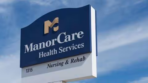 Blue ManorCare Health Services sign on a white post against a clear blue sky.